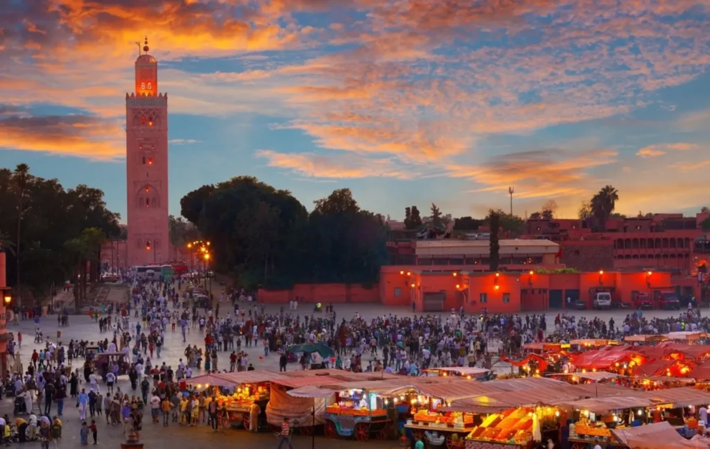 High-angle view of Jemaa el-Fnaa square in Marrakesh at sunset, featuring the Koutoubia Mosque minaret, glowing market stalls, and a bustling crowd under a vibrant orange sky, its a highlits of Morocco itinerary 5 days