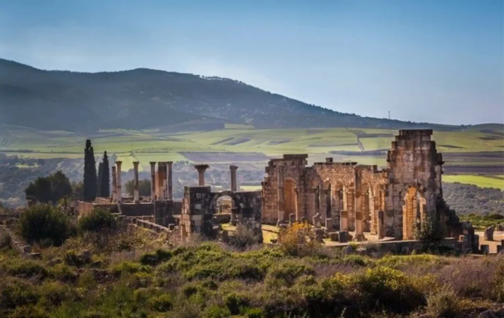 Morocco itineraries highlighting the ancient Roman ruins of Volubilis with stone columns and arches set against a backdrop of green hills and mountains.