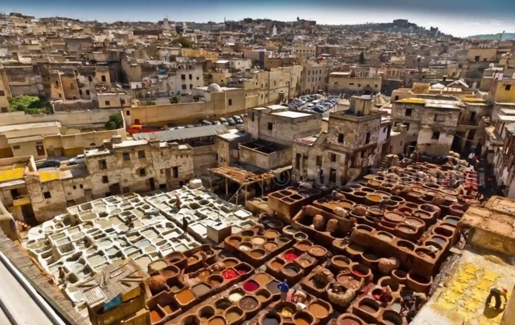 A high-angle view of the Chouara Tannery in Fes, showing dozens of round stone vats filled with colorful dyes and workers processing leather amidst the historic city buildings.