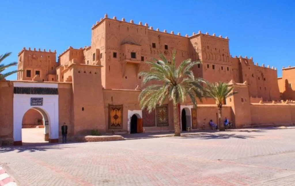 A large, ancient reddish-brown clay fortress with palm trees in front and a clear blue sky in Ouarzazate, Morocco.
