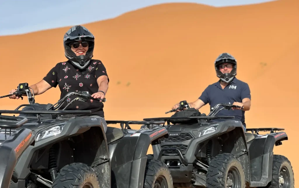 A man and a woman smiling while riding black ATVs on orange sand dunes under a clear blue sky in Morocco.