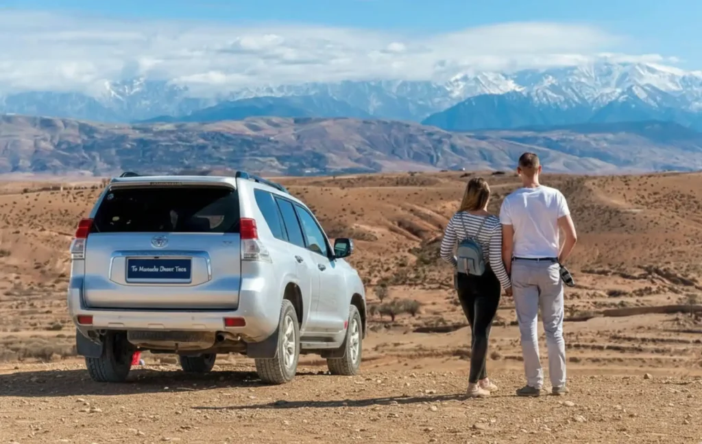 Morocco itineraries showing a couple standing next to a silver Toyota Land Cruiser looking out over the desert landscape toward the snow-capped Atlas Mountains.