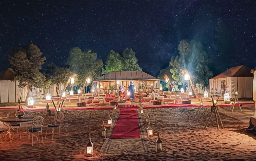 Morocco itineraries featuring a luxury desert camp at night with glowing lanterns, red carpets, and traditional tents under a starry sky in Merzouga.