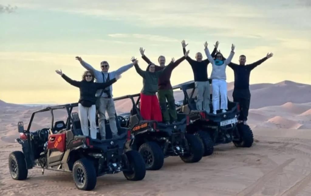 A happy group of travelers celebrating on their buggy Merzouga tour while standing on top of their off-road vehicles in the desert dunes.