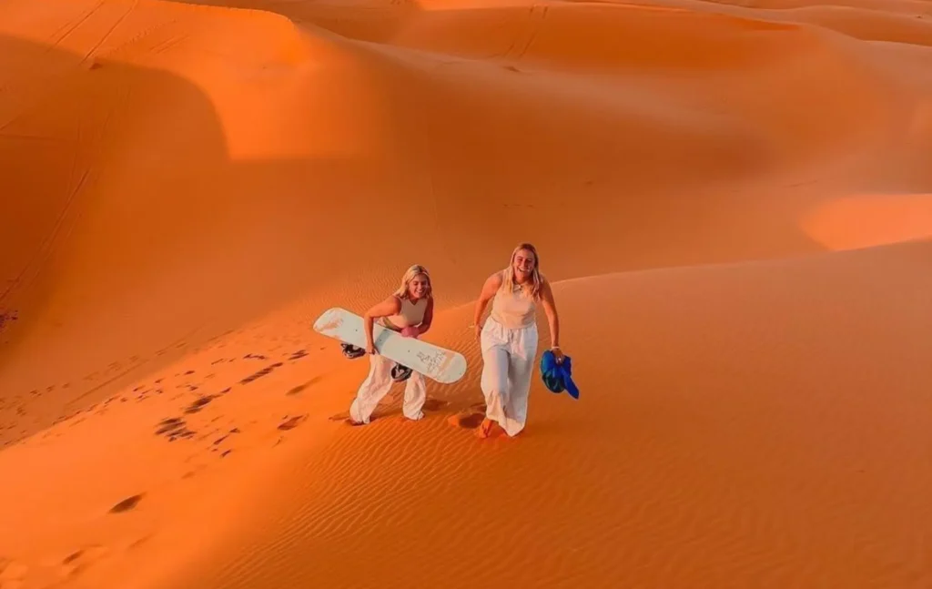 Two women walking up a golden Merzouga quad-style dune carrying sandboards at sunset.