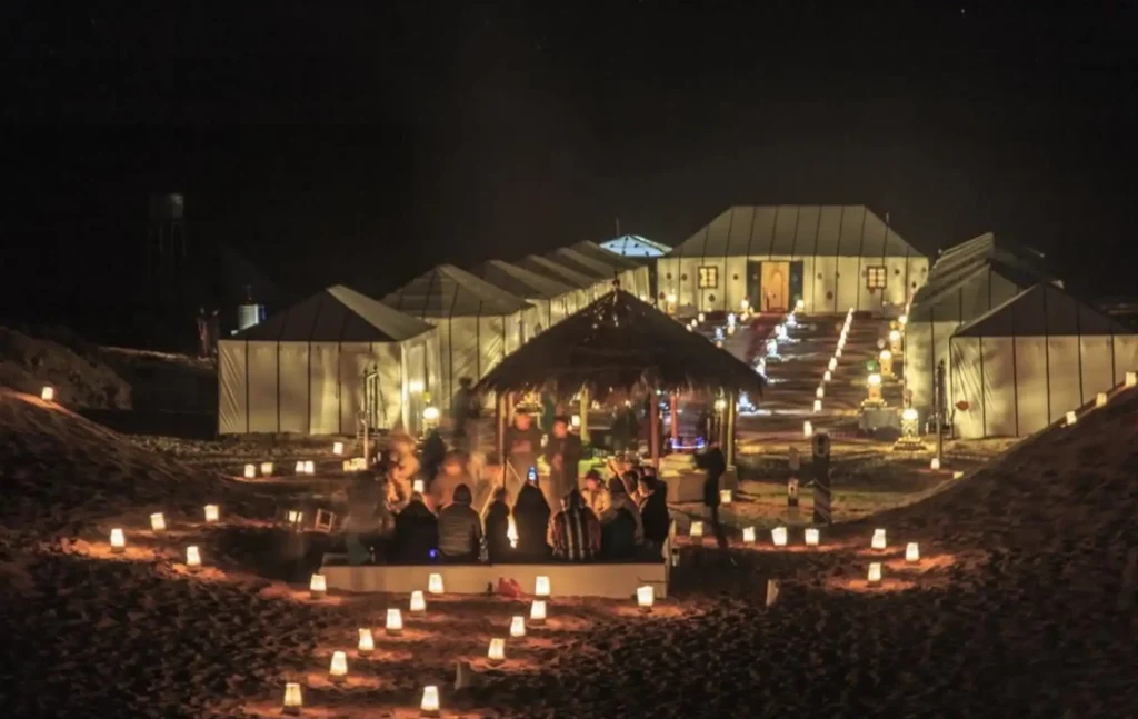 A glowing Merzouga luxury desert camp at night with lanterns illuminating the sand and guests gathered for Merzouga desert tours activities.