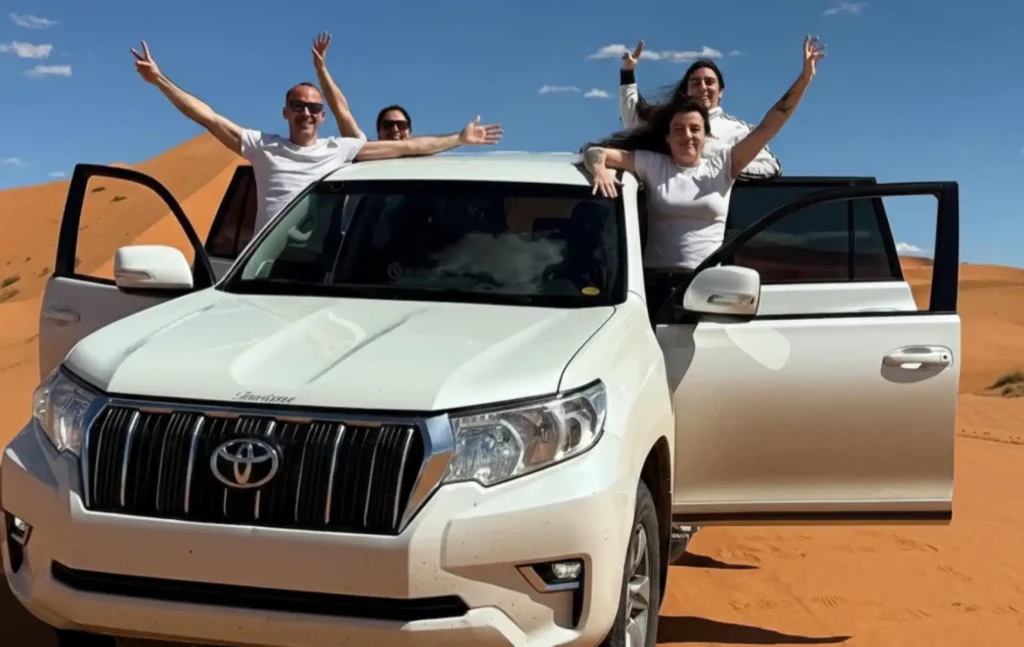 Happy travelers posing with a 4x4 vehicle in the sand dunes during a 4 day desert tour from Marrakech to fes.