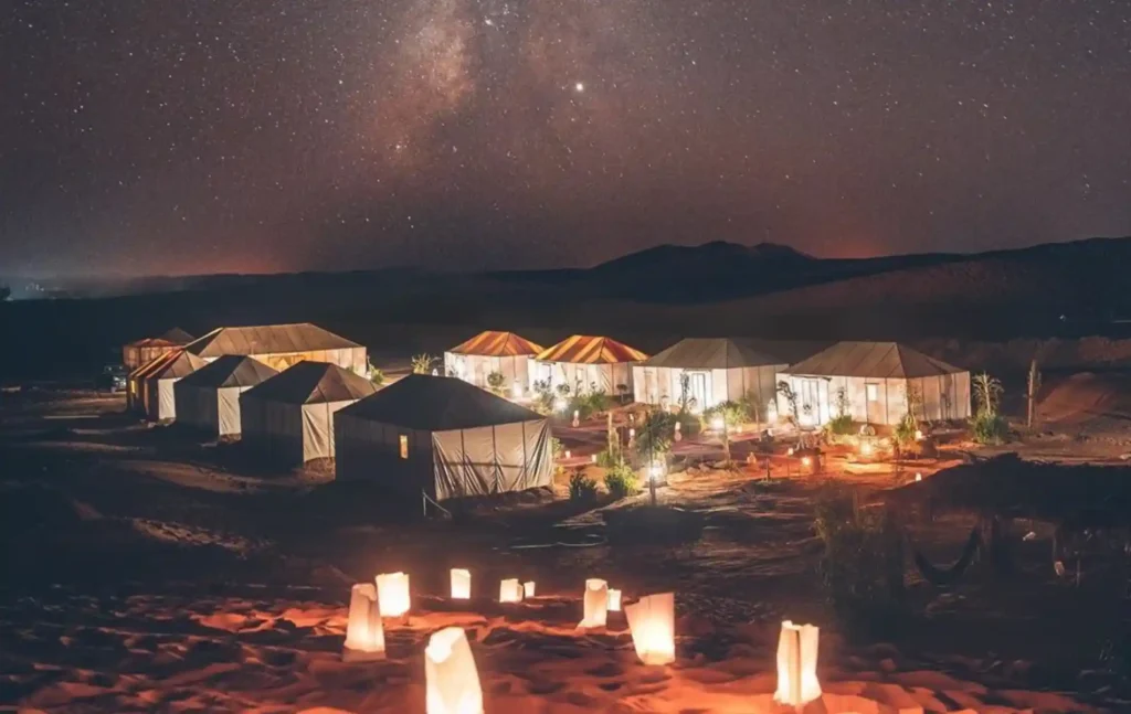 A beautiful luxury desert camp lit by lanterns under a clear Milky Way galaxy sky during a 4 days tour from Marrakech to Merzouga.
