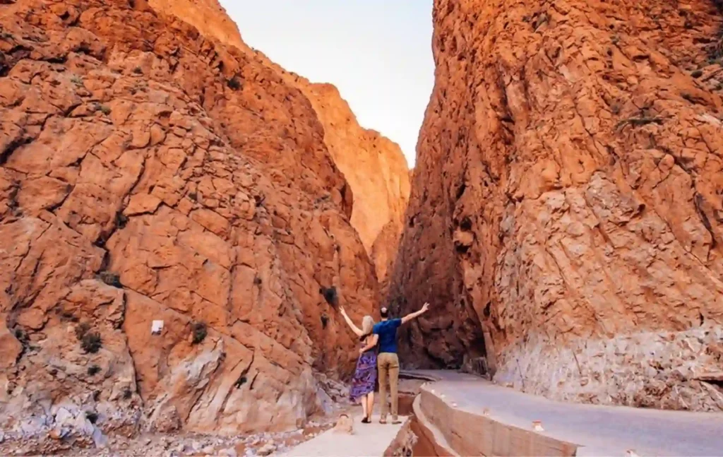 A couple standing with arms wide open between the massive red rock walls of the Todra Gorges during a 4 days tour from Marrakech to Merzouga.