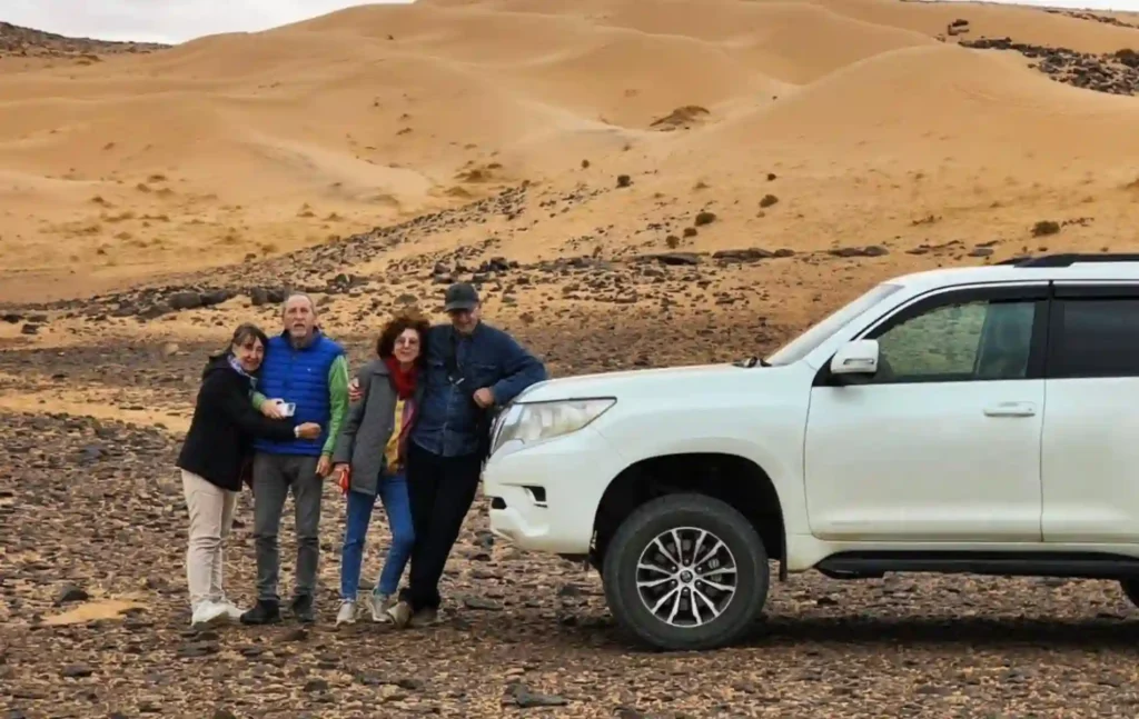 Four travelers posing next to a white Toyota Land Cruiser 4x4 in front of sand dunes during a 5 day desert tour from marrakech.