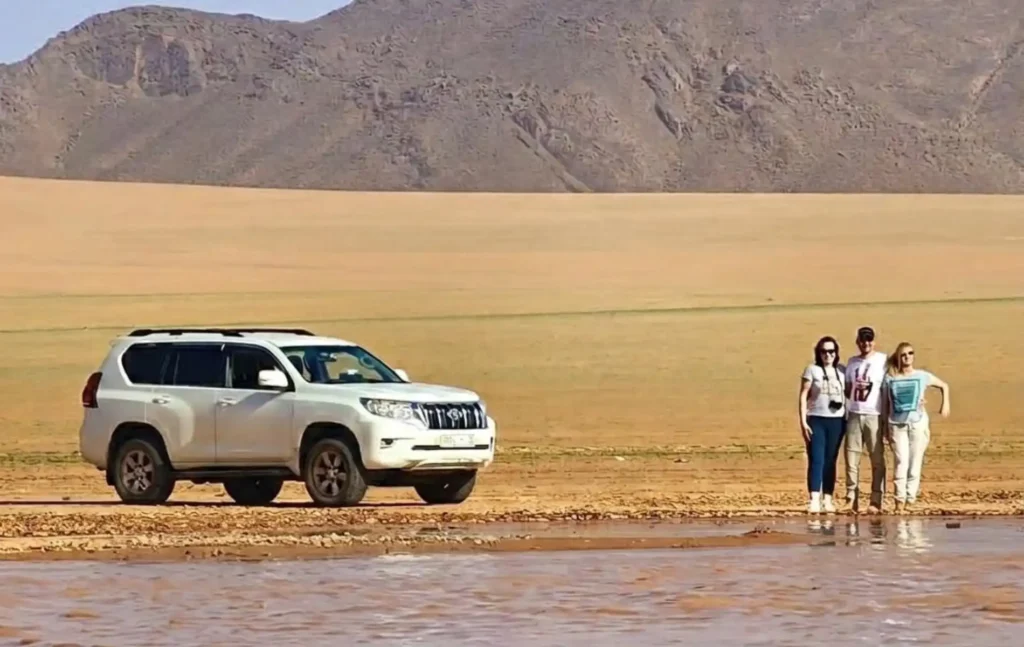 A white Toyota Prado 4x4 parked by a desert oasis in Tafraout Sidi Ali during a 5 day desert tour from marrakech with three travelers standing near a shallow river.