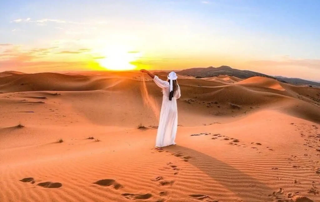 A woman in a white dress pouring sand from her hand during a marrakech to fes desert tour 2 days at sunrise.
