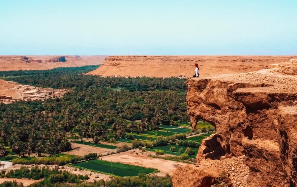 A traveler sitting on a high rocky cliff overlooking a lush palm grove and desert oasis during a 5 days tour from fes to marrakech.