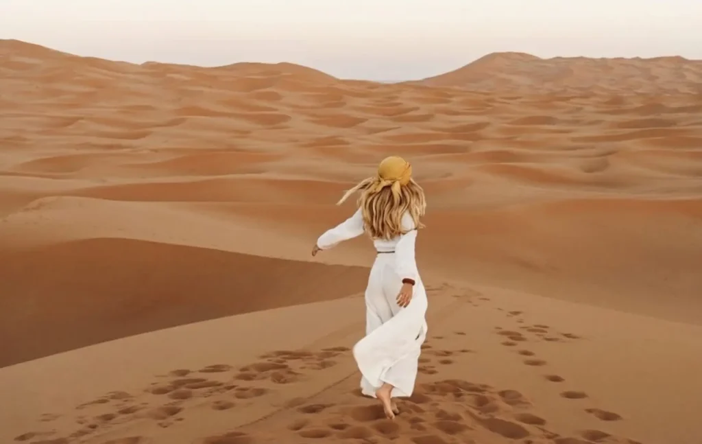 A woman in white walking across the sand dunes at sunrise during a 3 days desert tour from Fes to Marrakech.