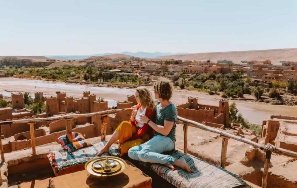 A couple sitting on a rooftop terrace enjoying traditional tea overlooking the ancient earthen buildings of Ait Ben Haddou during a 3 days desert tour from Fes to Marrakech.