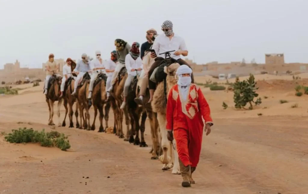 A line of travelers riding camels through the Moroccan desert led by a guide in traditional red clothing during a 3 days desert tour from Marrakech.