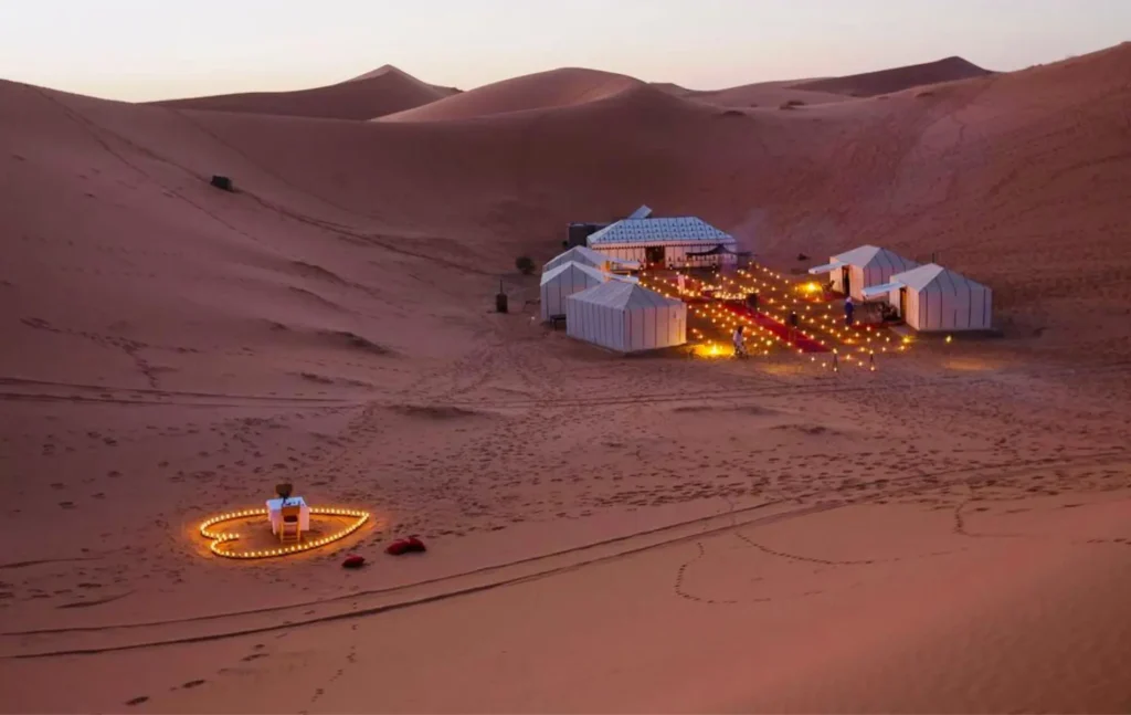 A luxury desert camp with white tents and a heart-shaped candle dinner setup in the dunes on a 2 days desert tour from fes.