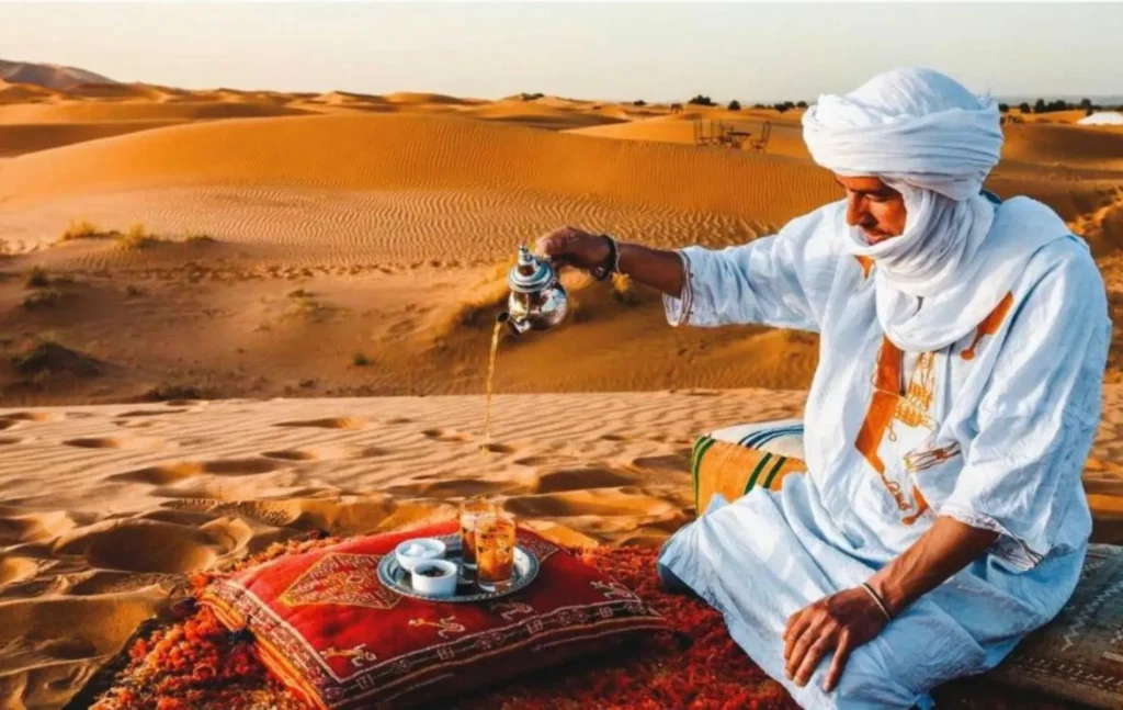 A Berber man in traditional blue clothing pouring Moroccan mint tea on a rug in the dunes during a 2 days desert tour from fes.