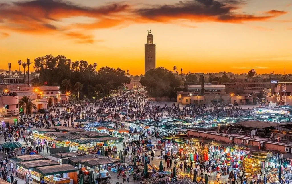 Morocco itineraries featuring the bustling Jemaa el-Fnaa square at sunset with market stalls, crowds, and the Koutoubia Mosque minaret in the distance.