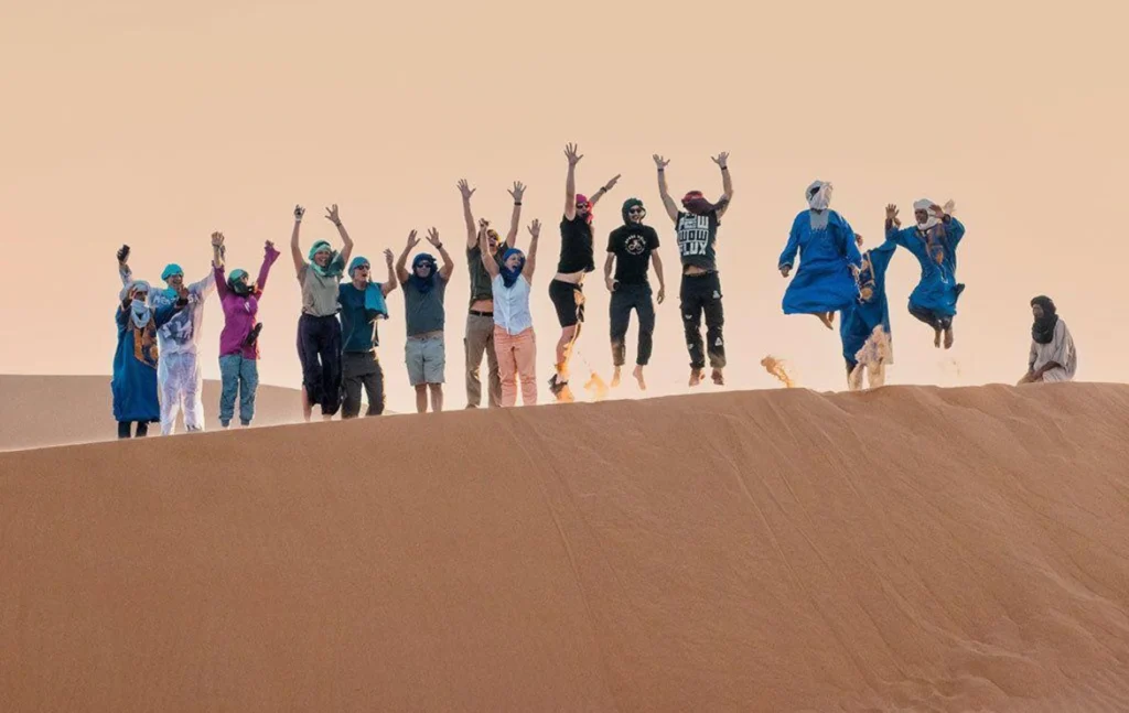 A happy group of tourists and guides jumping in the air on top of a sand dune during a shared marrakech to fes desert tour 3 days.