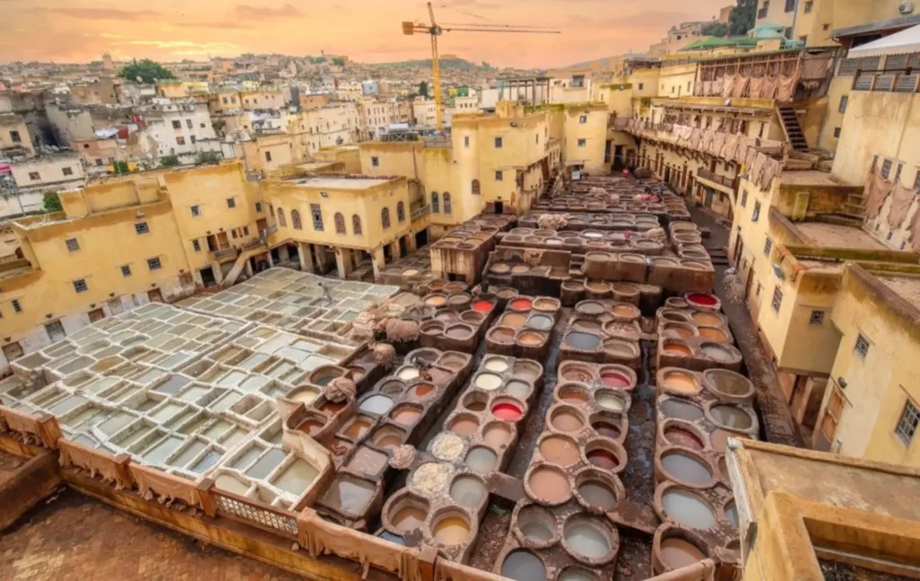 7 days morocco tour highlights the traditional leather tanning process at the famous Chouara Tannery in Fes where dozens of round stone pits are filled with colorful natural dyes surrounded by ancient yellow buildings under a soft sunset sky to showcase the authentic craftsmanship of the medina
