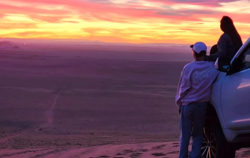 A traveler leaning against a white 4x4 vehicle watching a purple sunset over the desert during a 5 days desert tour from fse to marrakech with To Marrakech Desert
