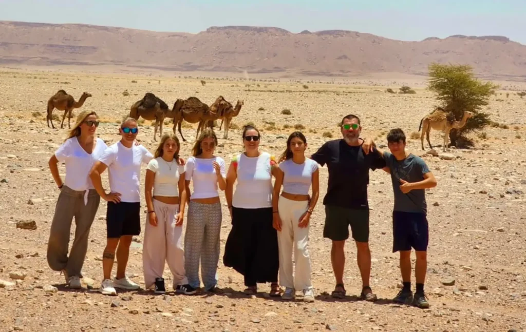 A group of diverse travelers standing together in the Moroccan desert with camels in the background during a 5 days desert tour from fes to marrakech.
