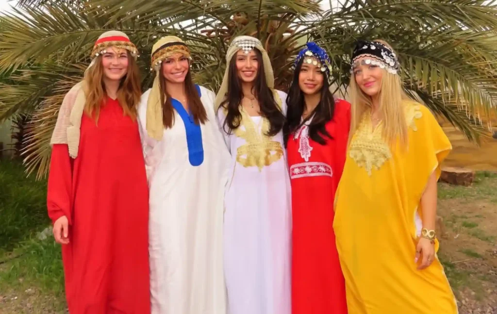 4 days morocco student trip: Five smiling female students wearing traditional Moroccan kaftans and ornate headpieces with coins while posing in front of a palm tree.