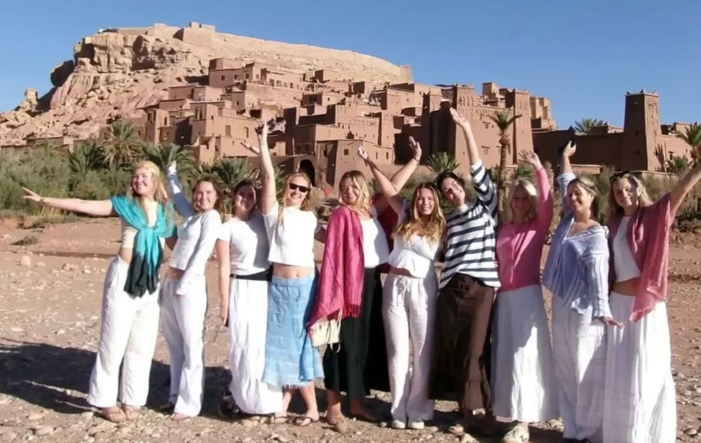 4 days morocco student trip: A large group of cheerful female students posing with arms raised in front of the historic earthen clay architecture of Ait Ben Haddou.