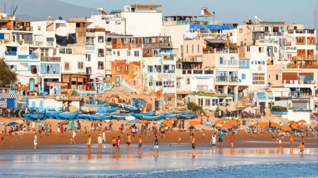 A busy sandy beach with crowds of people and blue fishing boats below a hillside village during Casablanca desert tours, an excellent coastal addition to a morocco itinerary.