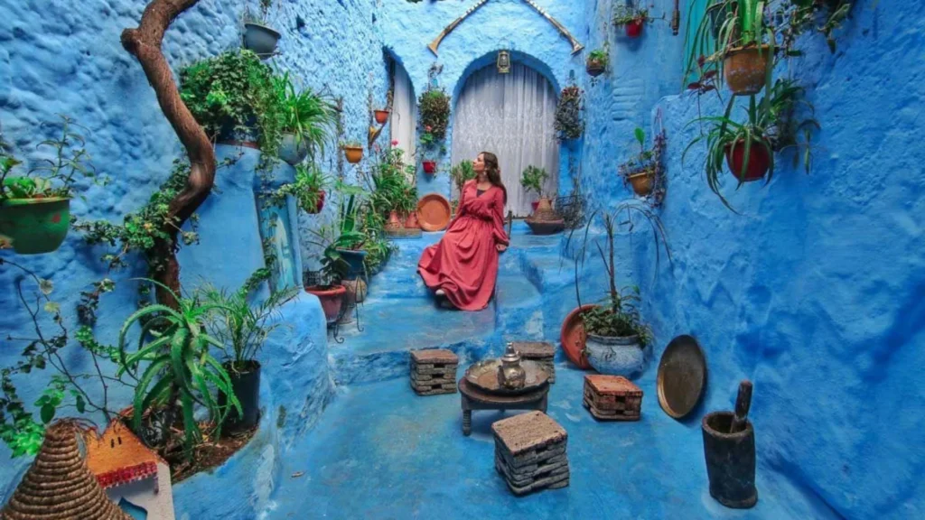 A woman in a long red dress sits on steps within a narrow, completely blue-washed alleyway in Chefchaouen decorated with potted plants, illustrating Tangier desert tours.