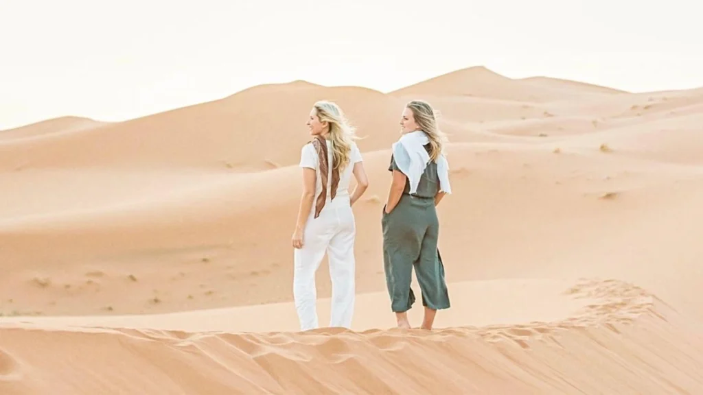 Two female travelers with scarves standing on the ridge of a golden sand dune looking out over the vast desert landscape, illustrating Tangier desert tours.