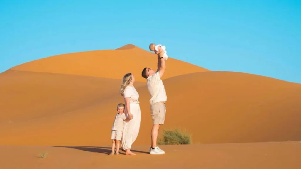 A family of four standing on a massive golden sand dune in the Sahara Desert under a clear bright blue sky, with the father joyfully lifting a baby into the air while the mother holds hands with their young son, illustrating our Marrakech Desert Tours.
