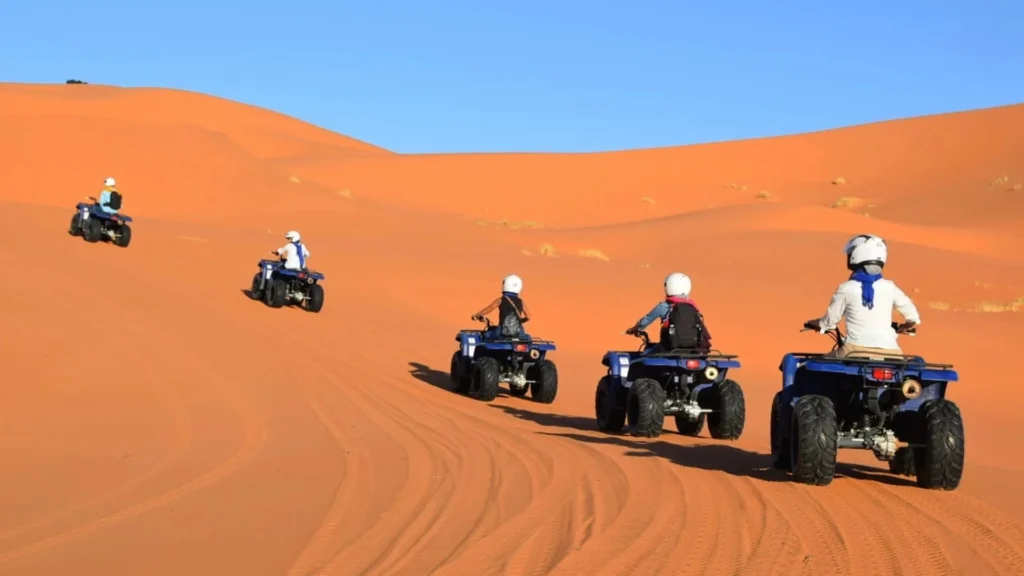 A line of travelers wearing safety helmets riding blue quad bikes in a row up a massive orange sand dune under a clear blue sky, illustrating our exciting Marrakech Desert Tours.