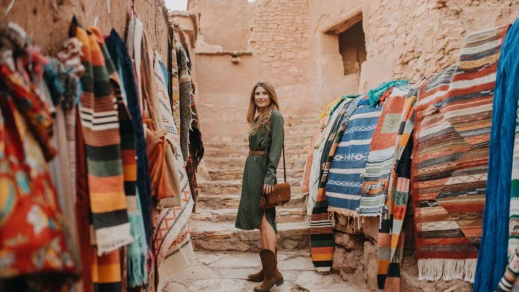 A woman walking through an ancient clay street lined with colorful carpets during Casablanca desert tours, an essential stop on a morocco itinerary.