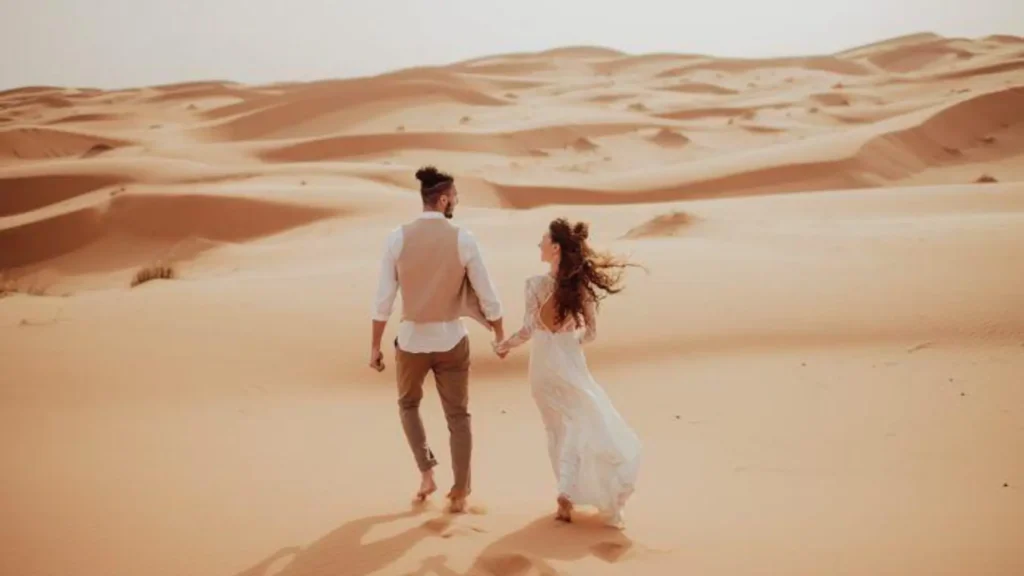 A romantic couple holding hands and walking barefoot on sand dunes during Casablanca desert tours, an unforgettable part of a morocco itinerary.