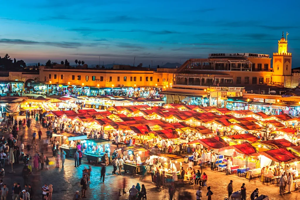 Is it Safe to Travel to Marrakech: A busy and brightly lit Jemaa el-Fnaa square at night filled with food stalls, locals, and tourists under a deep blue evening sky.
