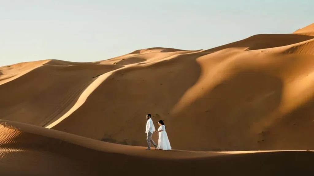 A romantic couple dressed in white clothes holding hands while walking along a sweeping sand dune ridge in the vast Sahara Desert under a clear sky, highlighting Marrakech desert tours.