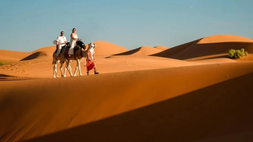 A wide landscape photo showing a couple dressed in white riding camels led by a local guide across smooth golden sand dunes under a clear sky, highlighting Marrakech desert tours.