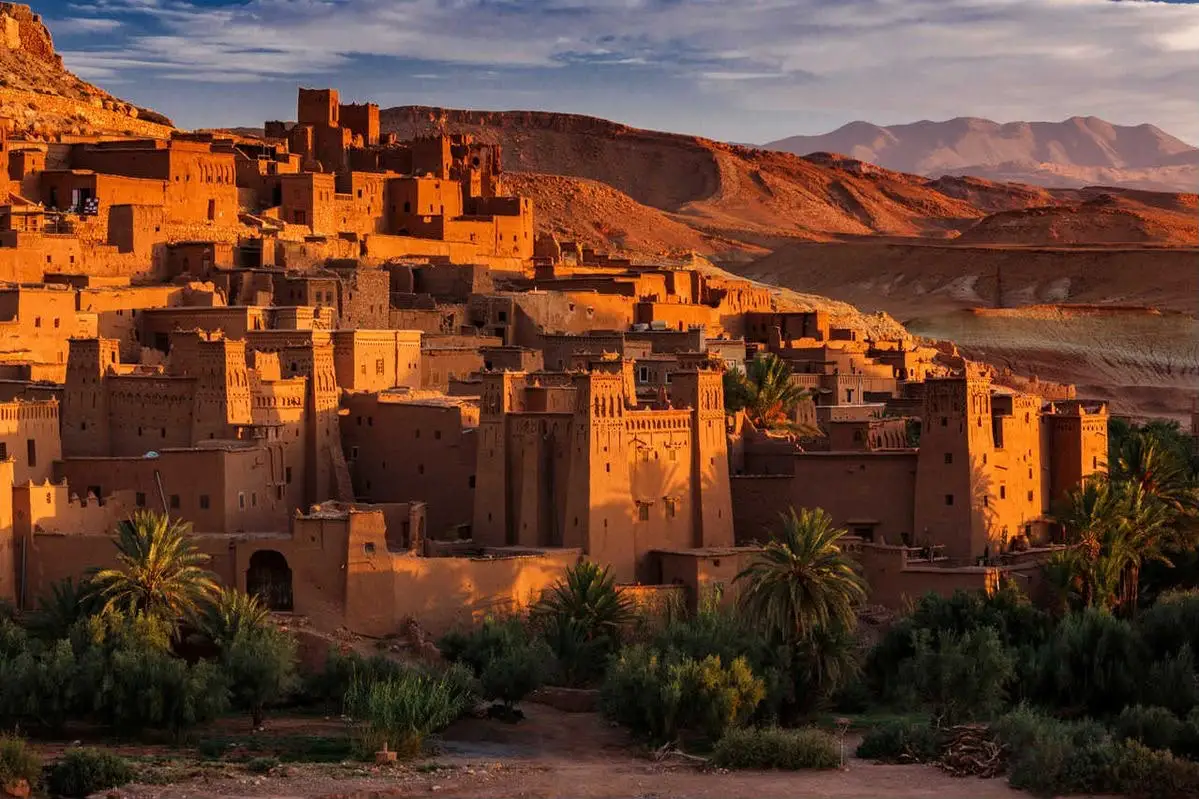 A breathtaking wide landscape view of the historic clay Kasbah of Ait Ben Haddou glowing orange under the warm golden light of a sunrise, with palm trees in the foreground and rolling desert mountains under a partly cloudy sky, highlighting a day trip from Marrakech to Ouarzazate and Ait Ben Haddou.