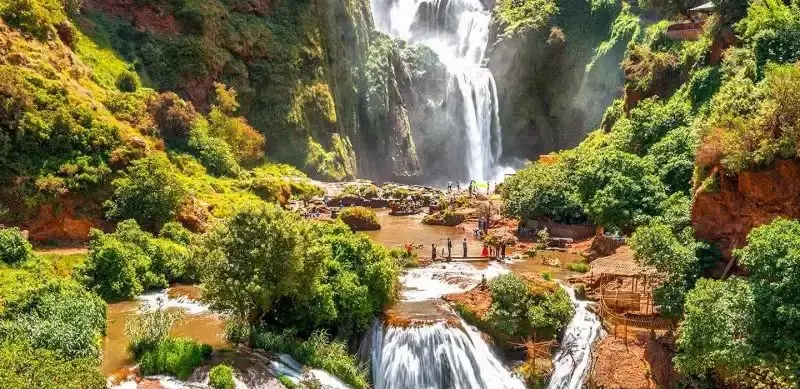 A vibrant wide landscape view of colorful cushions, patterned carpets, and low dining tables set up along the rocky banks of a rushing mountain river under bright sunlight, with a metal bridge and green valley slopes in the background, highlighting a day trip from Marrakech to Ourika Valley.