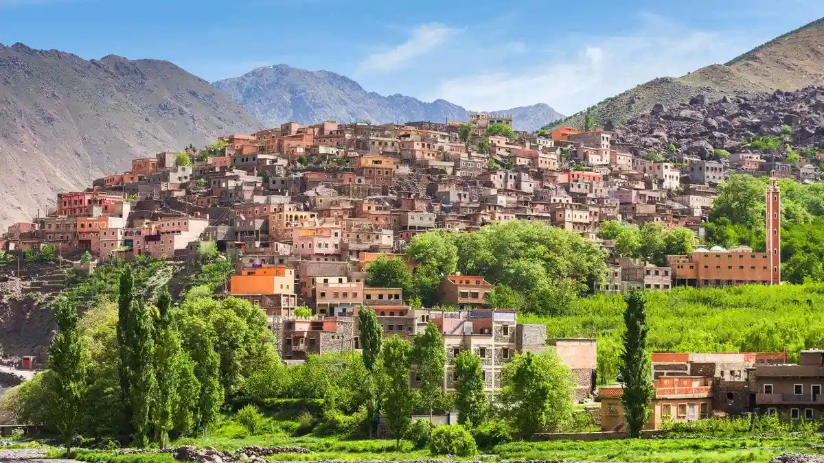 A scenic high-angle wide view of a traditional clay Berber village built into the slopes of the High Atlas Mountains, surrounded by lush green trees, terraced fields, and a prominent minaret under a bright blue sky, highlighting a day trip from Marrakech to Imlil.