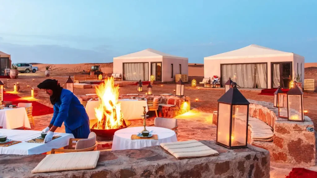 A staff member setting tables around a glowing campfire at a luxury desert camp in the Sahara, a highlight of Where to Visit in Morocco with To Marrakech Desert Tours.