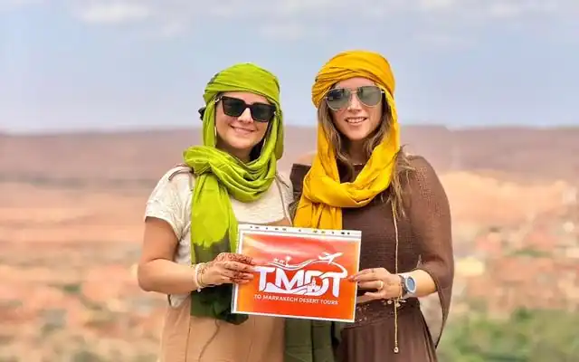 Two smiling travelers in colorful headscarves holding the To Marrakech Desert Tours sign